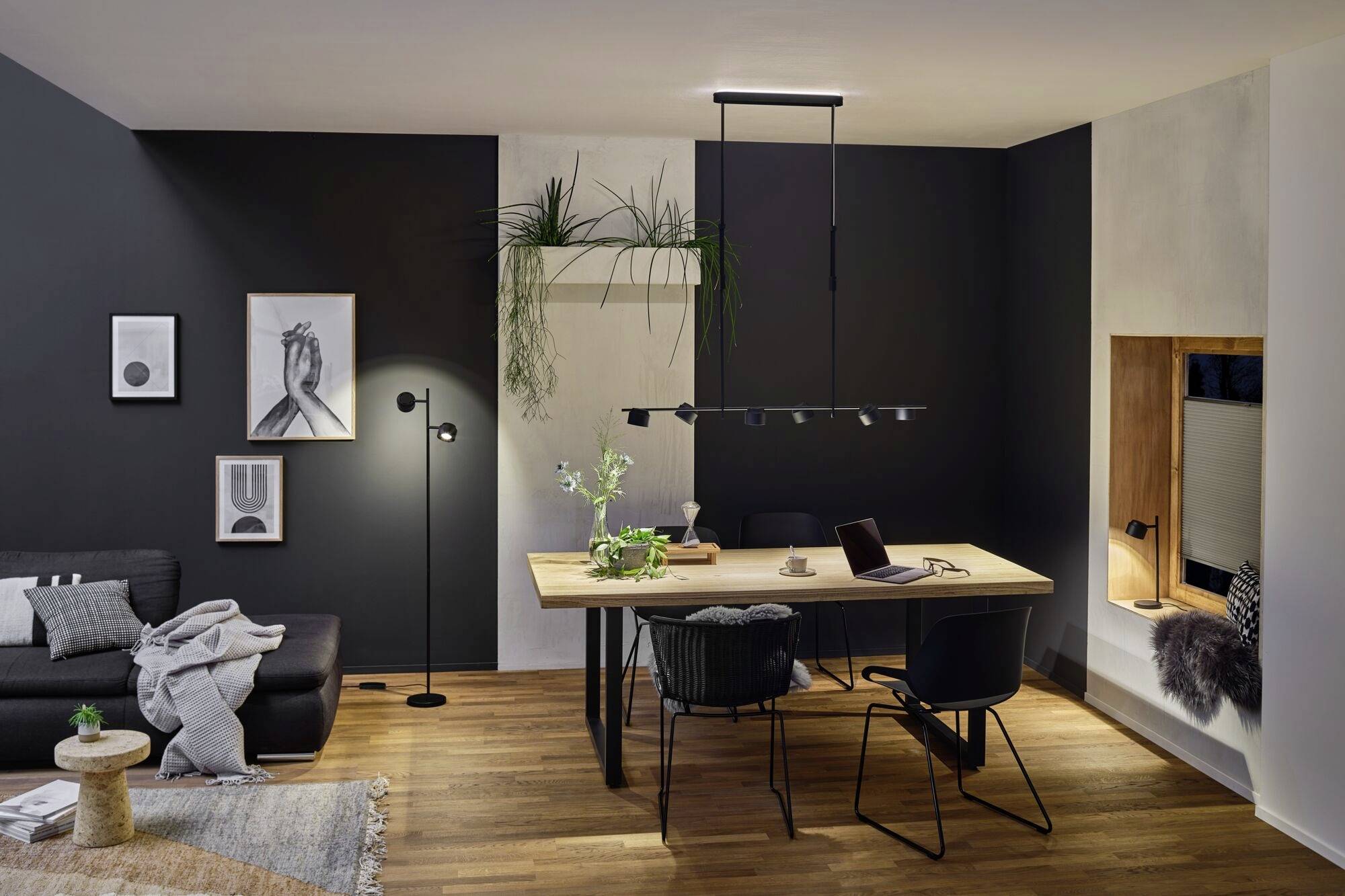 Esszimmer mit Holztisch, schwarzen Stühlen und moderner Beleuchtung. Pflanzen und Bilder schmücken die dunklen Wände.