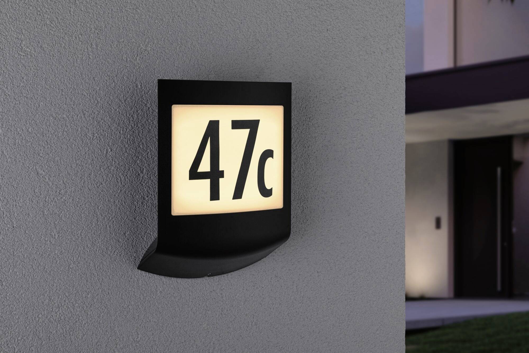 Hausnummernleuchte an einer Wand zeigt '47c'. Es ist Abend und die Lampe beleuchtet sanft die Umgebung.