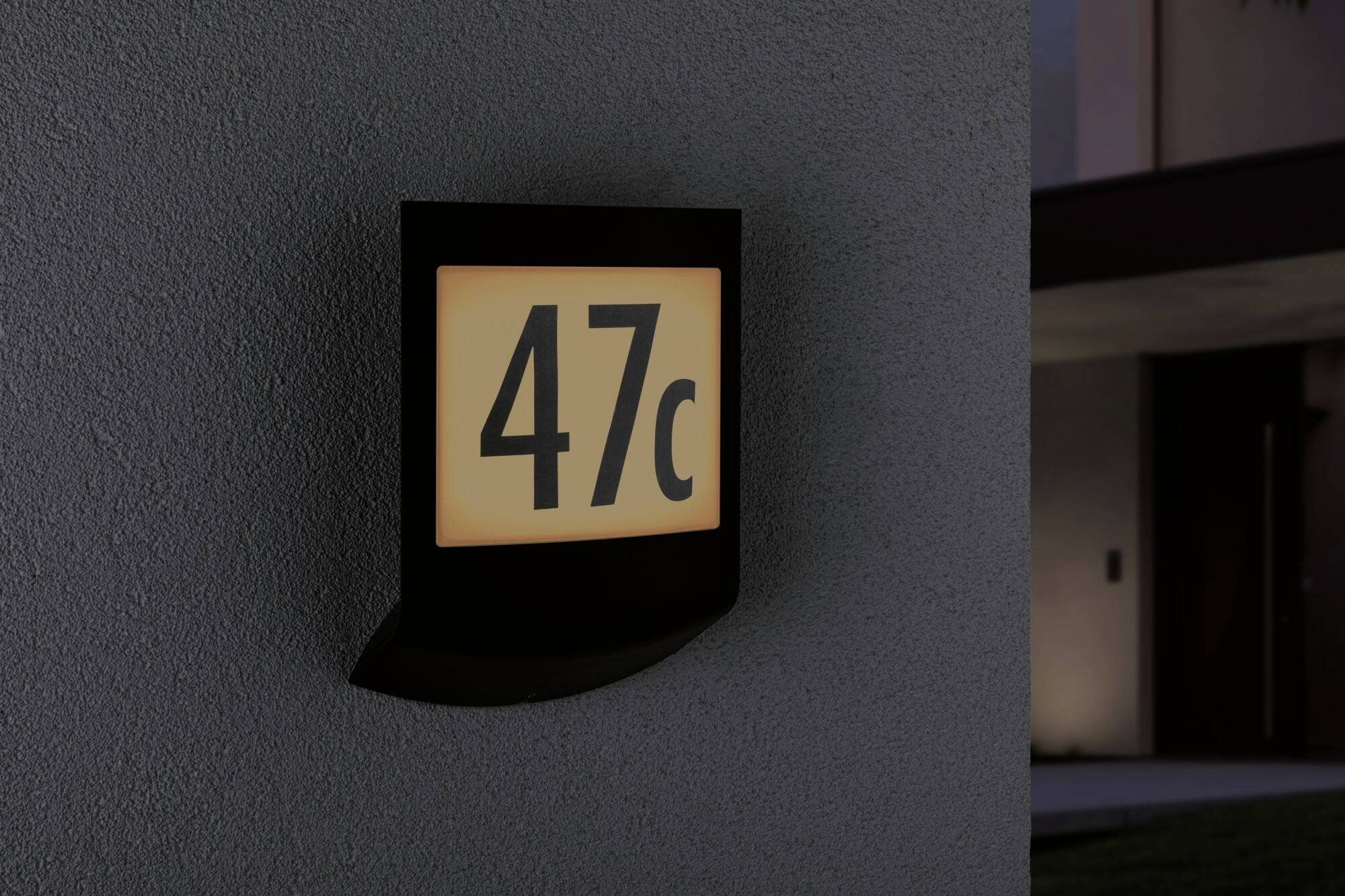 Beleuchtetes Hausnummernschild mit der Nummer '47c' an einer Außenwand, abends aufgenommen.
