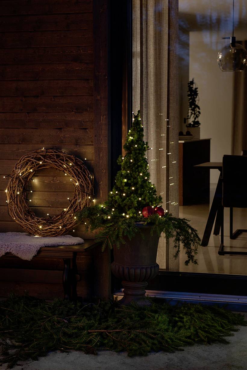Noch ein weihnachtlich dekorierter Eingang mit einem leuchtenden Kranz an der Wand. Ein kleiner Baum im Topf ist mit Lichtern geschmückt.