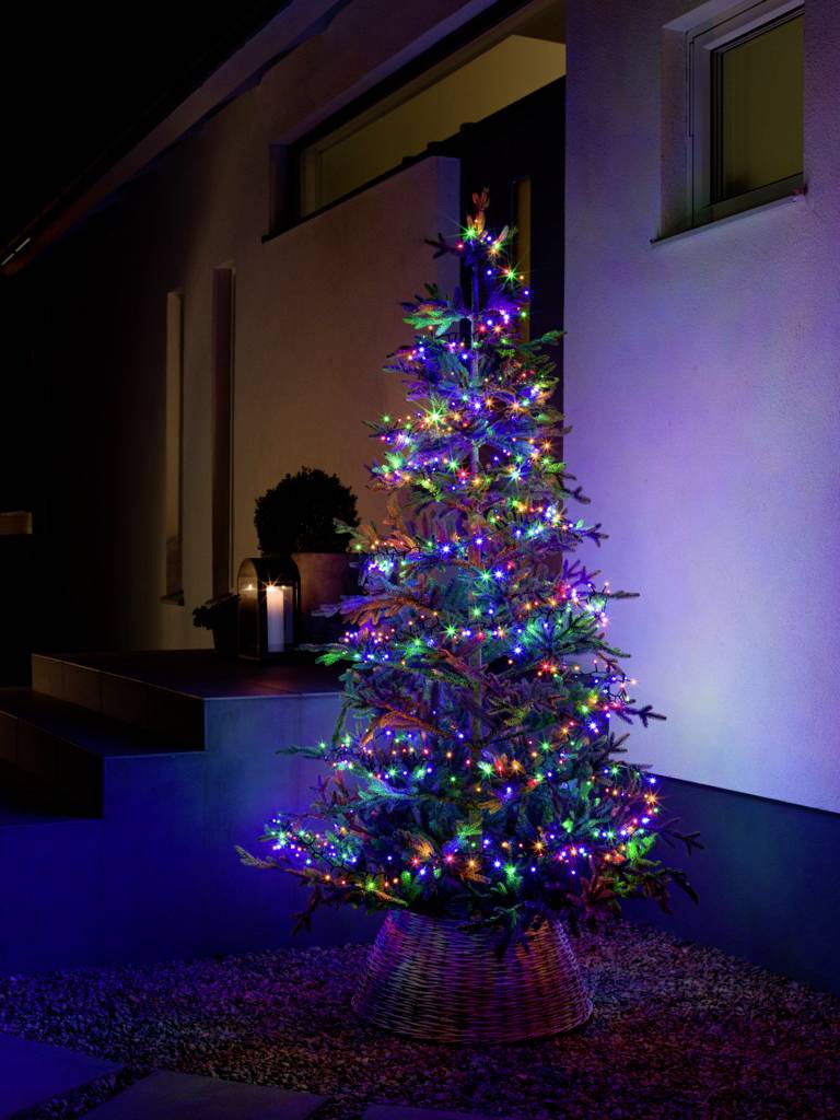 Ein Weihnachtsbaum im Freien, geschmückt mit bunten Lichtern, steht neben einem modern beleuchteten Hauseingang in der Nacht.