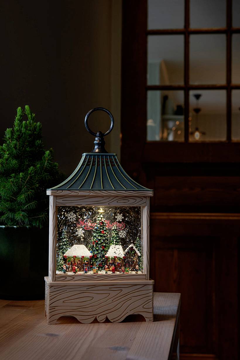 Weihnachtliche Laterne mit Miniaturdorf und bunten Lichtern auf einem Holztisch neben einem kleinen Tannenbaum, im Hintergrund unscharf.