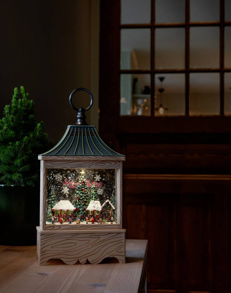 Eine festliche Laterne mit einer Miniaturszene aus Schnee und Tannen im Inneren steht auf einem Tisch neben einem kleinen Tannenbaum.