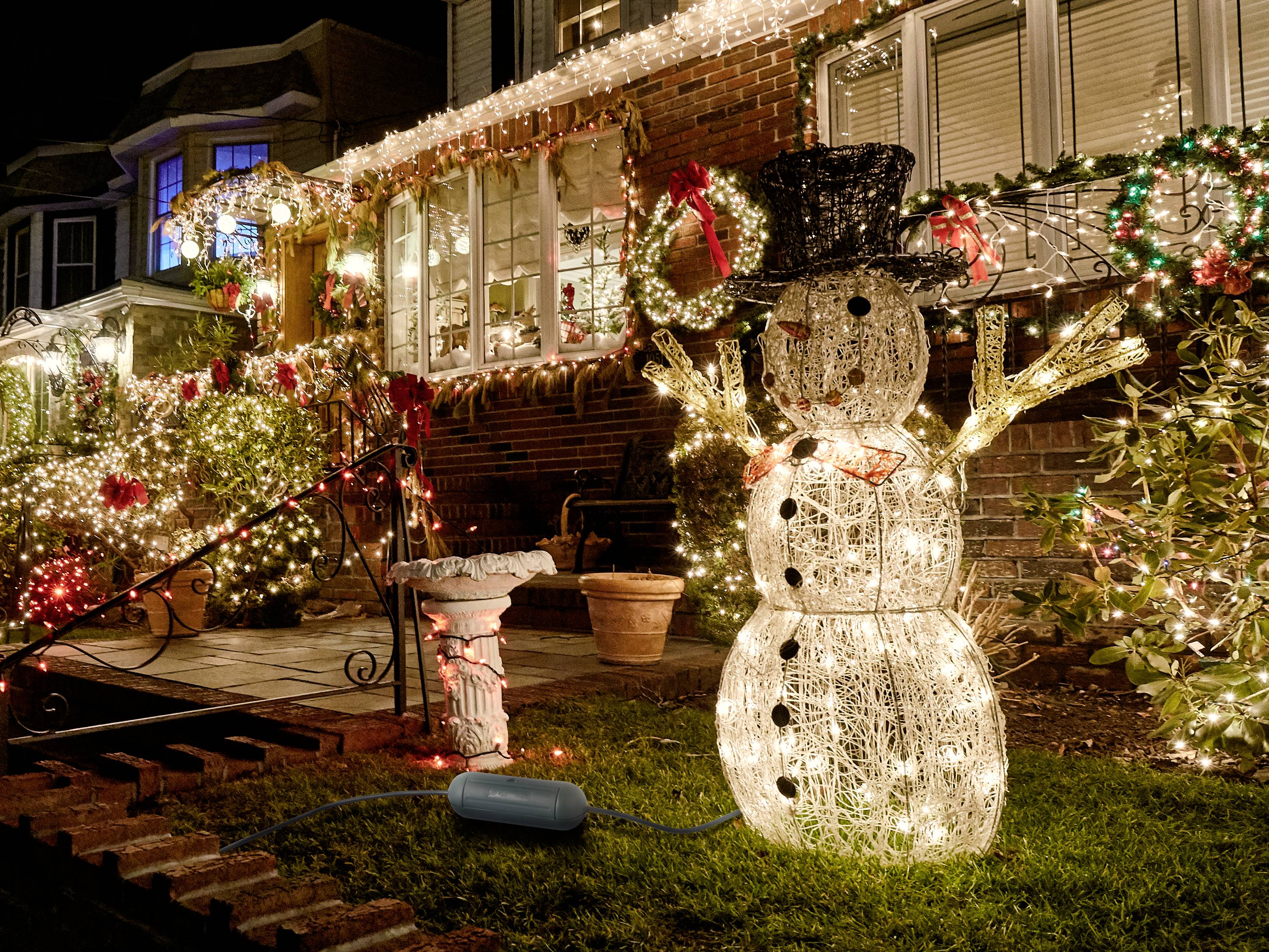 Ein beleuchteter Schneemann steht vor einem festlich geschmückten Haus mit Weihnachtsbeleuchtung und Dekorationen im Garten.