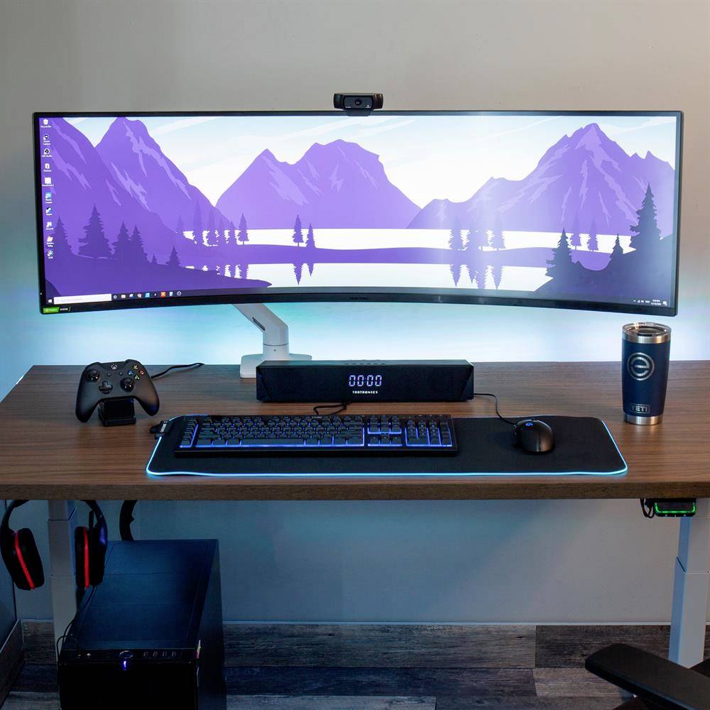 Ein moderner Schreibtisch mit einem gebogenen Monitor, auf dem eine Landschaft mit Bergen und See zu sehen ist. Controller, Lautsprecher und Tasse neben Tastatur.