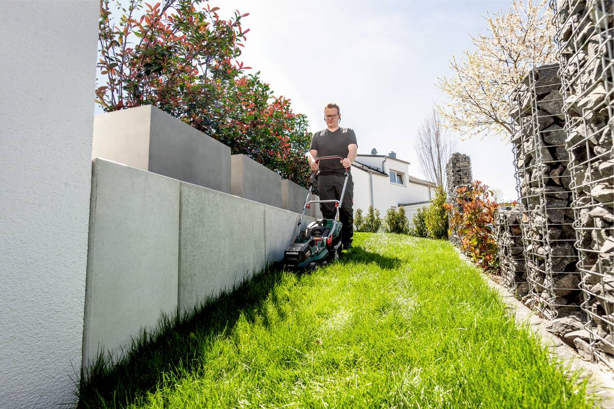 Eine Person mäht mit einem Rasenmäher einen kleinen, gepflegten Garten zwischen modernen Mauern und Blumentöpfen bei sonnigem Wetter.
