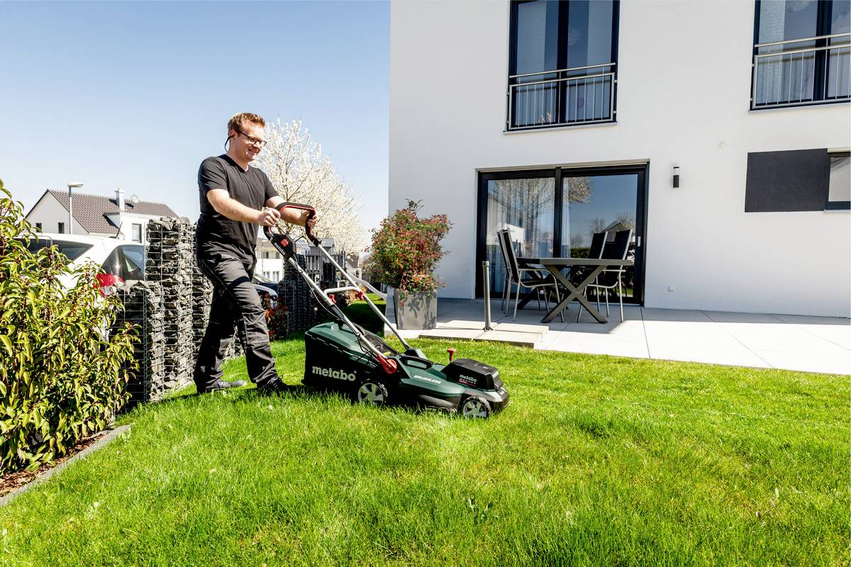 Eine Person mäht mit einem Rasenmäher Rasen im Garten vor einem modernen Wohnhaus.