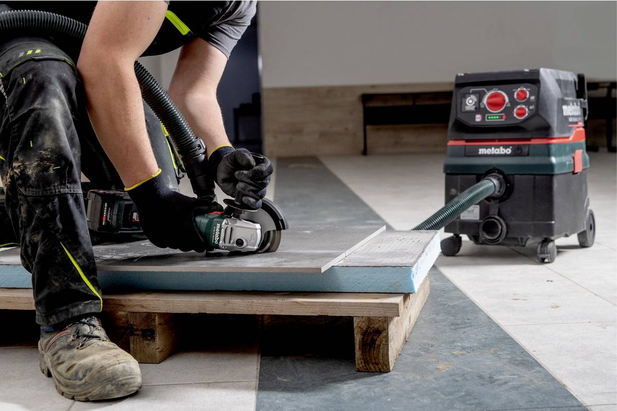 Arbeiter verwendet Elektrowerkzeug zum Schneiden von Baumaterialien auf einer Baustelle. Staubsauger saugt Staub nebenbei auf.