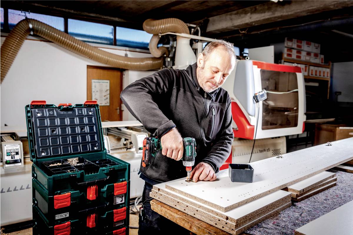 Ein Mann arbeitet in einer Werkstatt mit einer Bohrmaschine an einem Holzstück. Im Hintergrund steht eine Maschine und Werkzeugkisten.