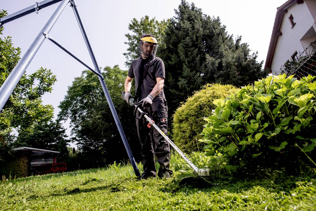 Eine Person mäht mit einem Rasentrimmer den Rasen in einem Garten. Umgeben von Büschen und Bäumen, trägt sie Schutzkleidung und einen Helm.