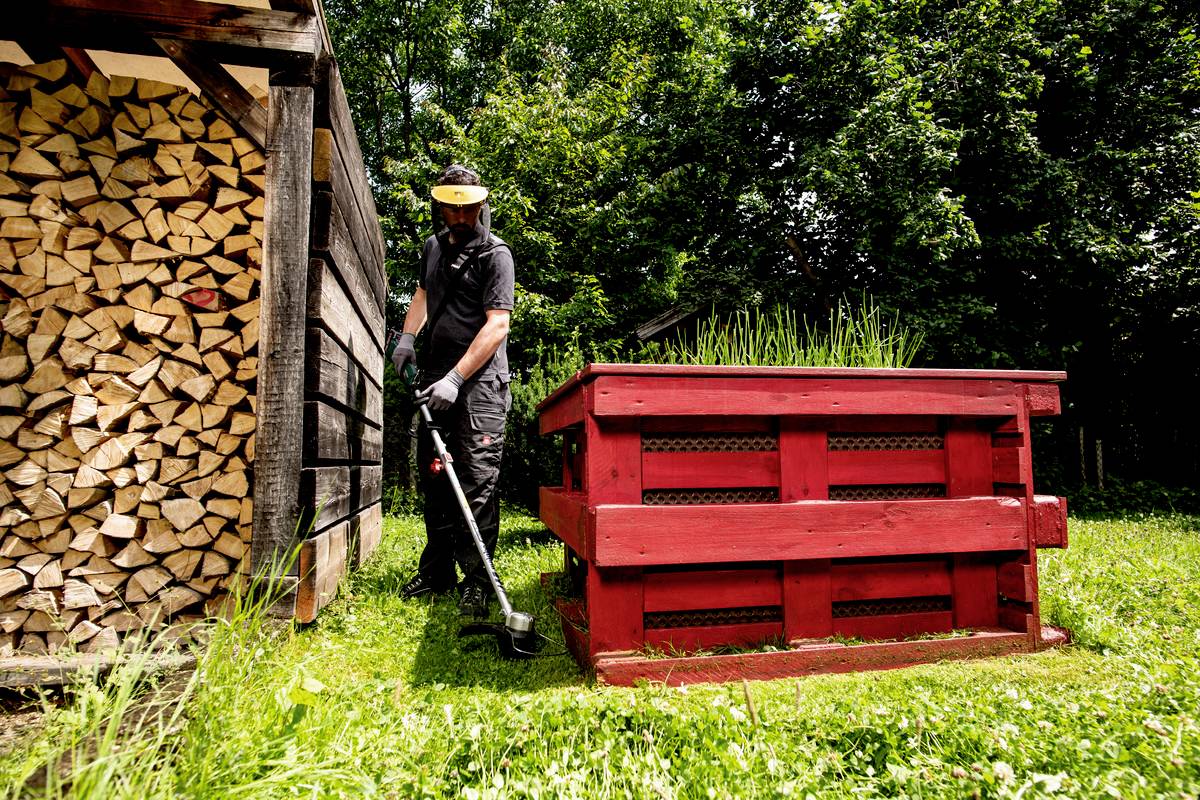 Eine Person mäht mit einem Rasentrimmer das Gras neben einem Holzstapel und einem roten Hochbeet. Im Hintergrund sind Bäume zu sehen.
