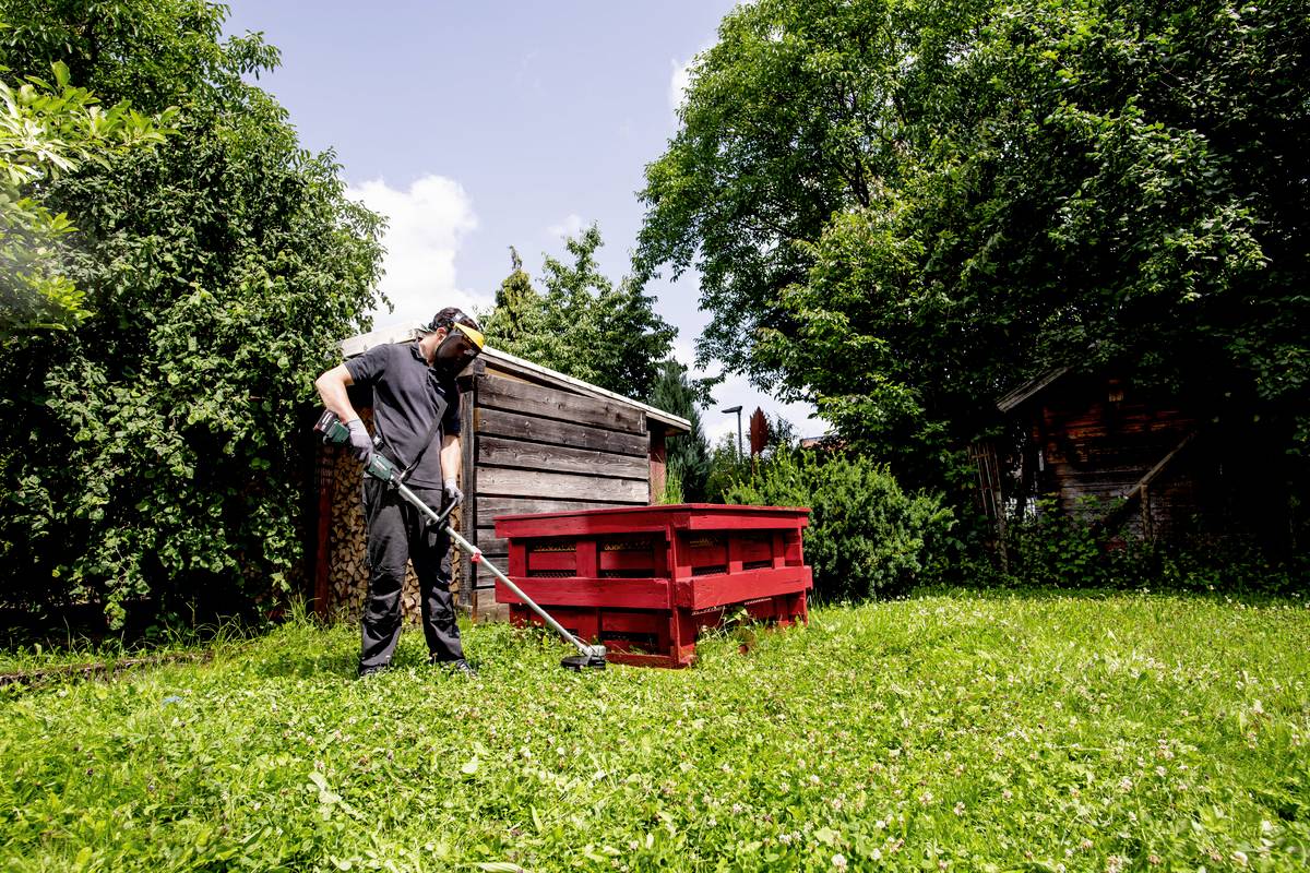 Eine Person mit Schutzbrille und Kopfhörern mäht das Gras in einem grünen Garten. Im Hintergrund sind rote Holzpaletten zu sehen.