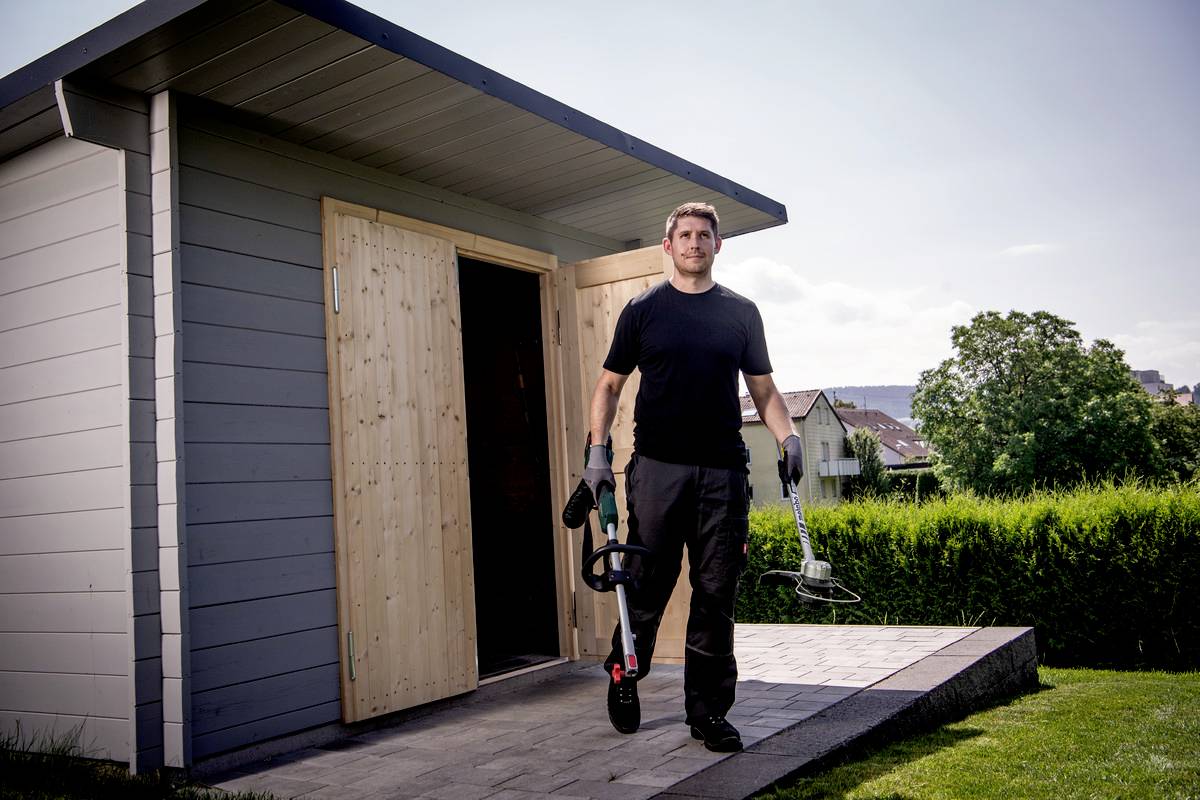 Ein Mann in Arbeitskleidung trägt Gartenwerkzeuge vor einem Geräteschuppen im Garten.