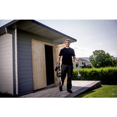 Ein Mann in Arbeitskleidung trägt Gartenwerkzeuge vor einem Geräteschuppen im Garten.