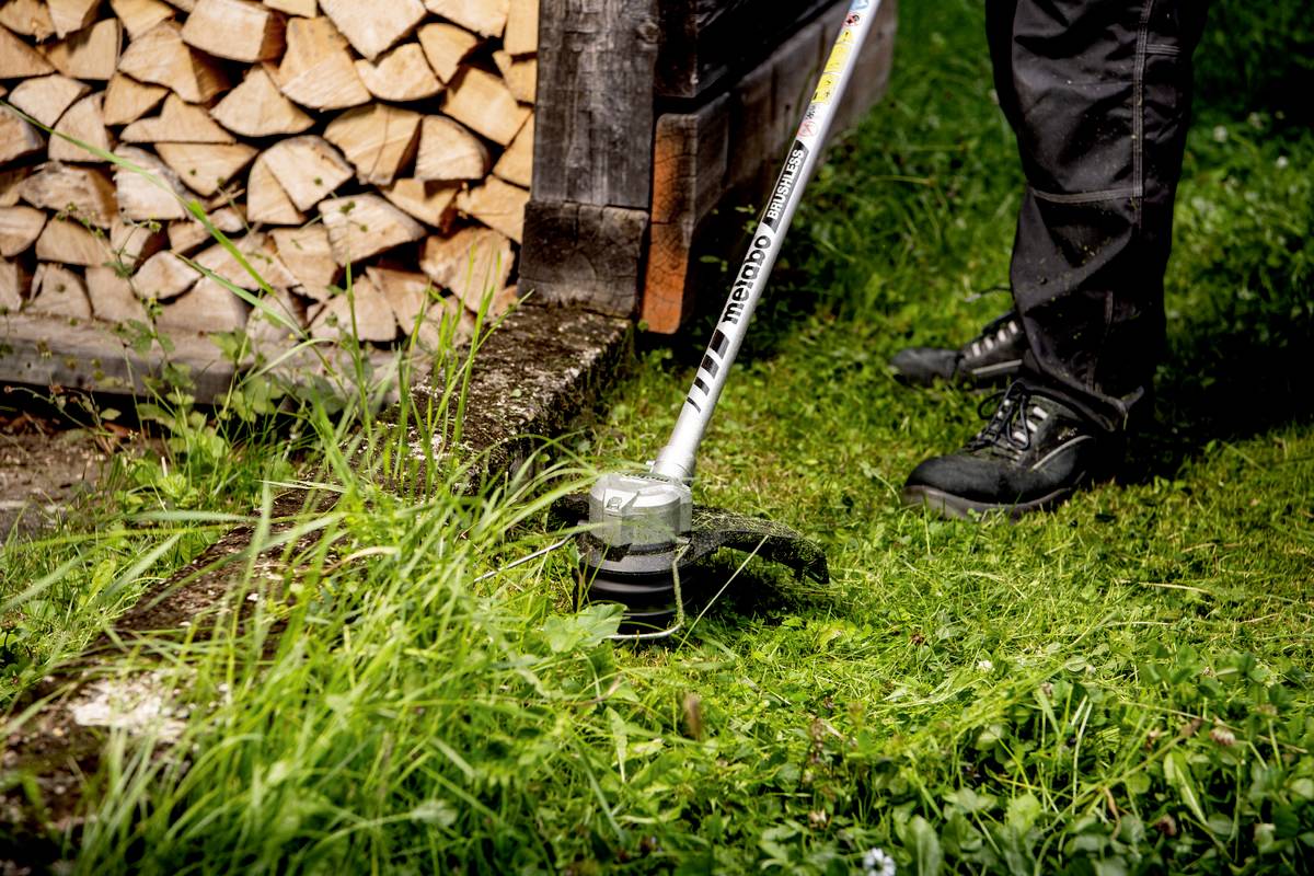 Eine Person benutzt einen Rasentrimmer, um Gras neben einem Stapel Feuerholz zu schneiden. Die Person trägt schwarze Schuhe.