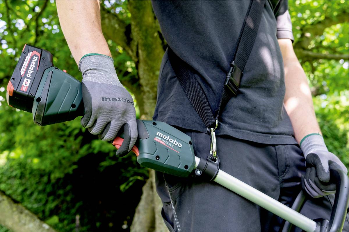 Eine Person hält eine grüne Akku-Gartenschere mit einem Metabo-Logo in einem grünen Garten. Trägt dunkle Kleidung und Handschuhe.