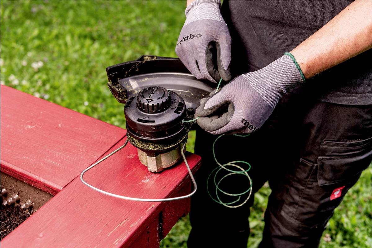 Eine Person mit grauen Handschuhen wechselt die Schnur in einem Rasentrimmer im Freien. Ein rotes Möbelstück ist sichtbar.
