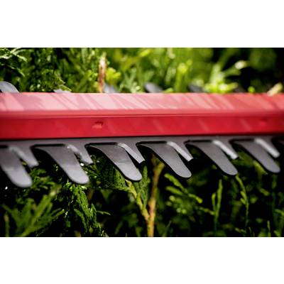 Eine rote Heckenschere schneidet grünes Laub einer Hecke. Die schwarzen Zähne der Schere sind deutlich sichtbar.