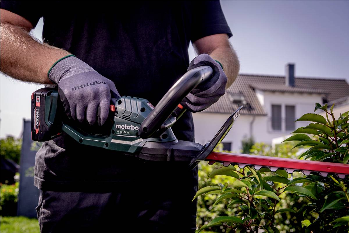 Eine Person schneidet Hecken mit einem elektrischen Heckenschneider im Garten. Häuser sind im Hintergrund zu sehen.
