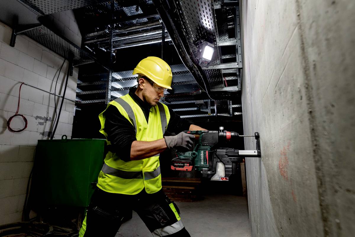 Ein Bauarbeiter mit Schutzhelm und Warnweste bohrt ein Loch in eine Betonwand, umgeben von Metallstrukturen und Werkzeugen in einem industriellen Raum.