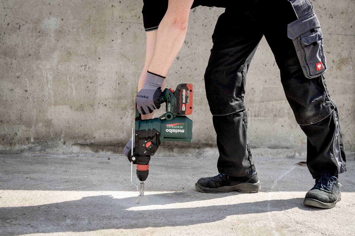 Person, die auf einer Baustelle steht und mit einem grünen Bohrhammer in den Boden bohrt. Trägt Arbeitshose und Handschuhe.
