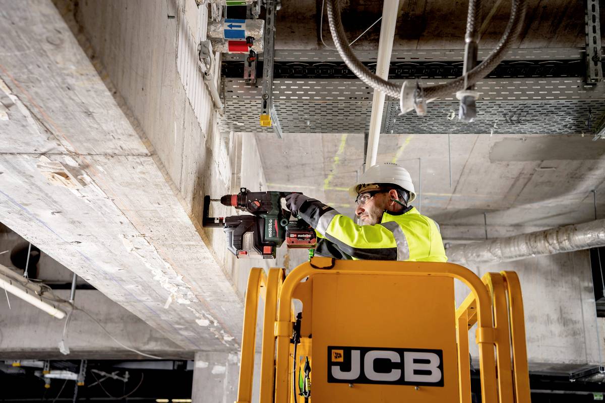Ein Bauarbeiter in Schutzkleidung arbeitet auf einer Hebebühne an einer Decke. Er benutzt ein Elektrowerkzeug zur Installation oder Reparatur.