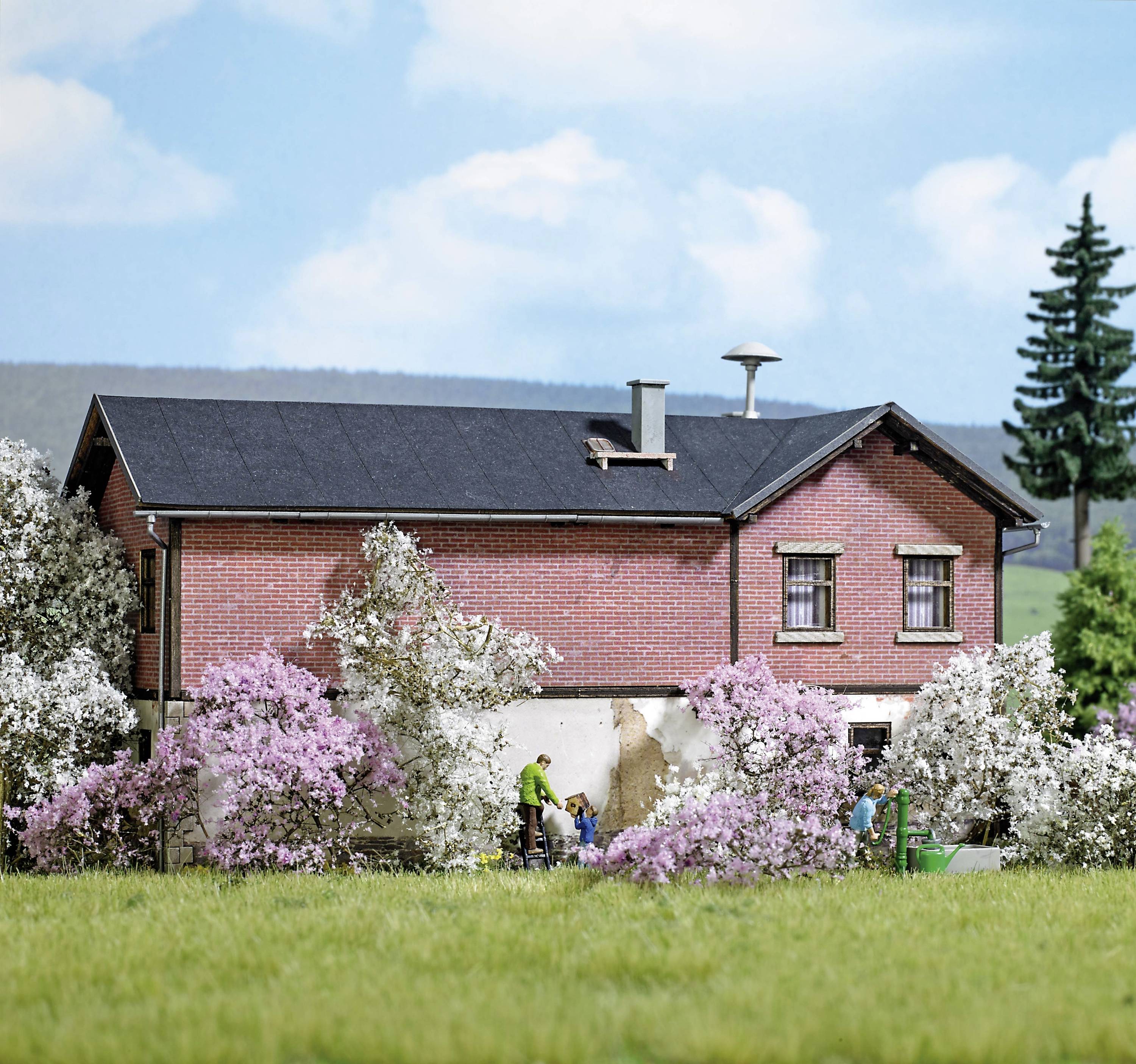 Ein rotes Backsteinhaus mit schwarzem Dach, umgeben von blühenden Bäumen. Personen arbeiten im Garten. Blauer Himmel im Hintergrund.