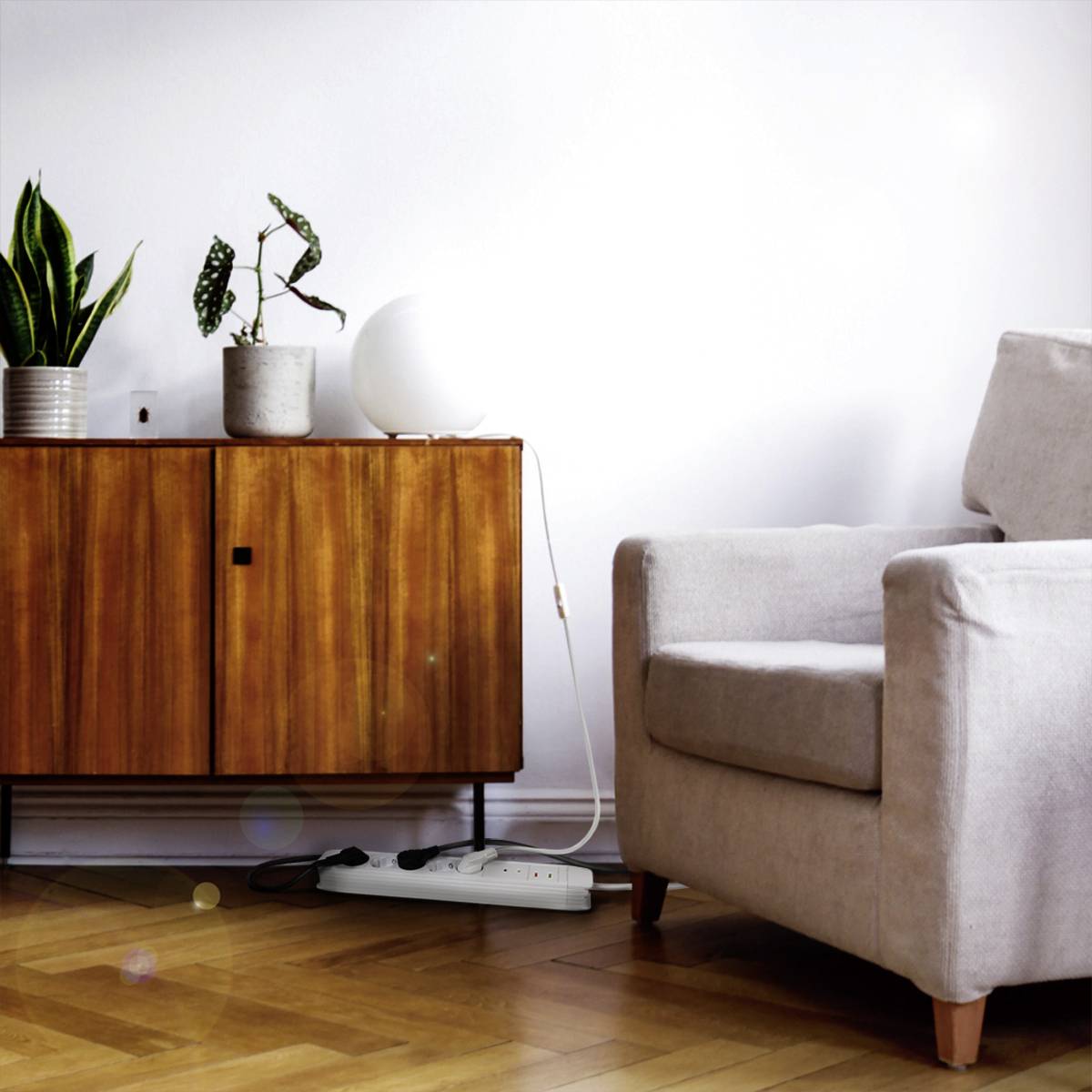 Ein Wohnzimmer mit einem grauen Sessel rechts, einem hölzernen Sideboard links mit Pflanzen darauf und einem Bodensteckdosenleisten davor.