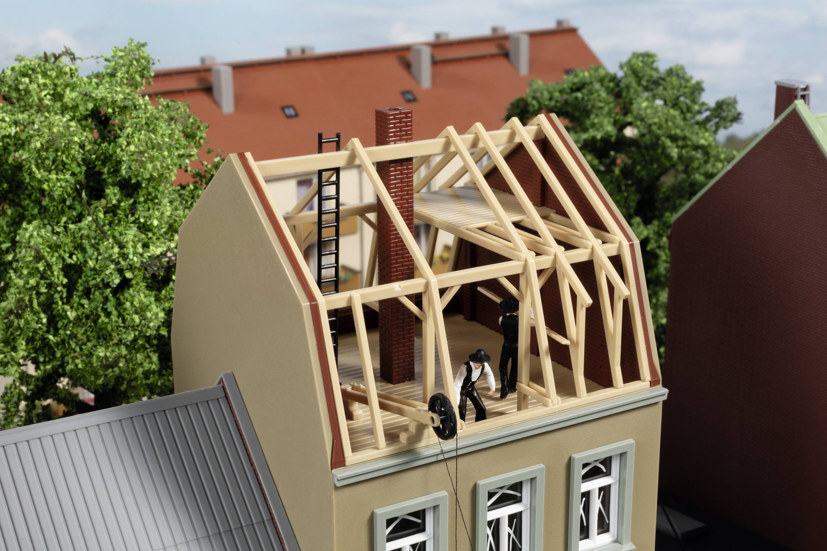 Ein Miniaturhaus zeigt einen Dachstuhlbau mit Holzbalken. Eine Figur steht auf dem Dach, hält ein Seil. Hintergrund: Bäume, Gebäude.