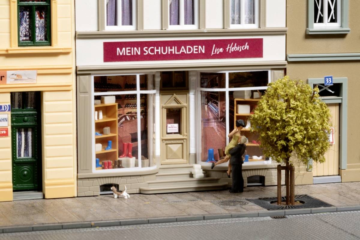 Ein Miniaturmodell eines Schuhgeschäfts mit der Aufschrift 'Mein Schuhladen'. Eine Person mit Kind betrachtet die Auslage.