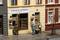 'Drei Figuren vor einem Geschäft mit dem Schild "Tabak & Whisky". Eine Person zeigt auf eine Ware auf einem Fass. Stadtstraße im Hintergrund.'