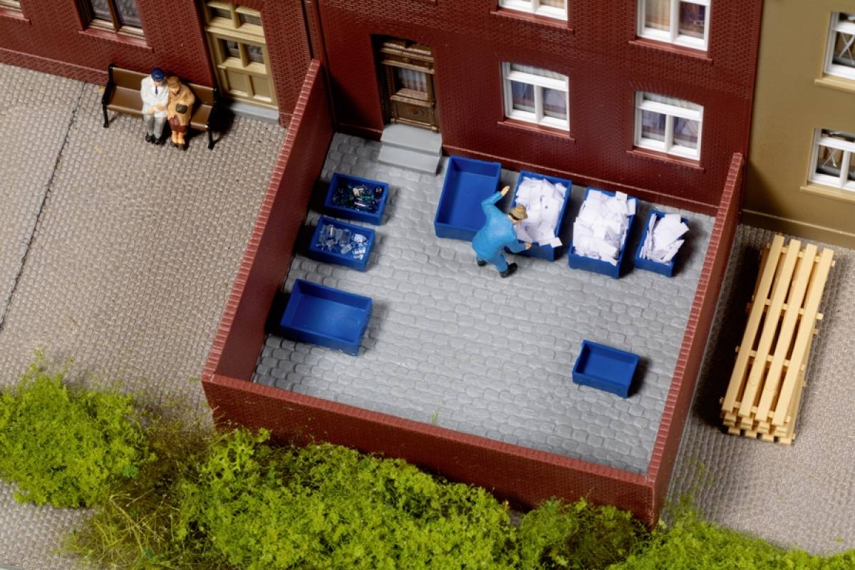 Eine Person recycelt Papier in blauem Müllbehälter im Hinterhof eines Wohngebäudes, daneben stapeln sich weitere Behälter. Kleines Modell.