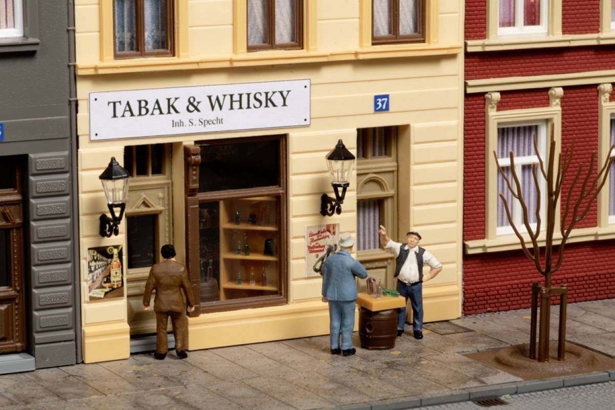 Drei Figuren stehen vor einem Tabak- und Whiskygeschäft. Ein Mann trägt eine Kiste, während ein anderer daneben steht. Ein Schild zeigt 'Tabak & Whisky'.