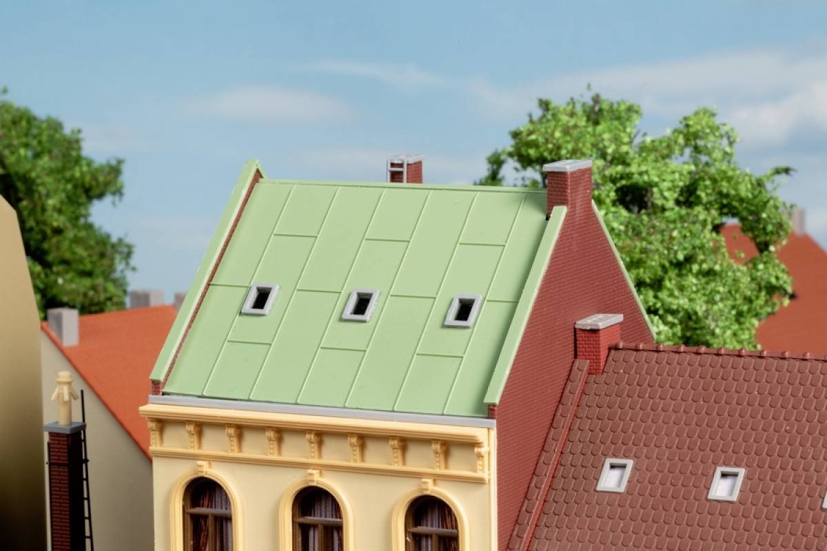 Ein Modell eines Altbaus mit grünem Dach. Im Hintergrund Bäume und Dächer weiterer Gebäude. Klarer Himmel über der Szene.
