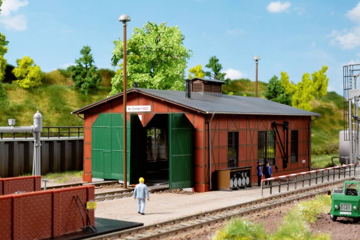 Ein kleines Eisenbahndepot neben Bahngleisen, umgeben von Bäumen und einer Wiese. Ein Arbeiter läuft in Richtung des Gebäudes.