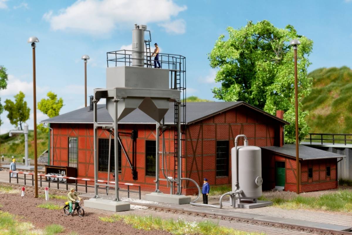 Eine Modelleisenbahnszene zeigt ein rotes Gebäude mit einem Wasserturm daneben. Zwei Figuren stehen am Gleis, eine Fahrradfahrerin fährt vorbei.