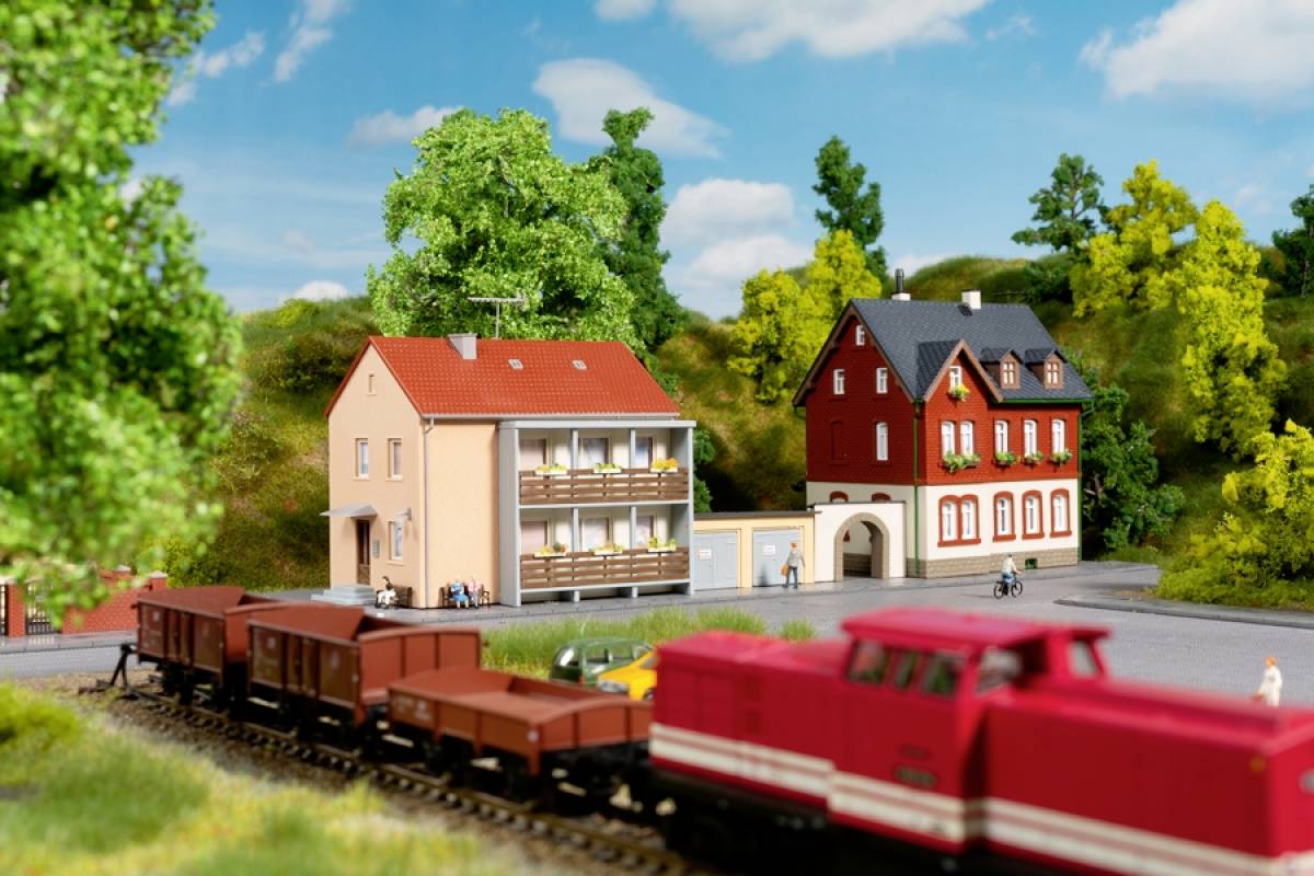 Eine rote Modelllokomotive fährt an einer kleinen Landschaft vorbei, mit zwei Häusern und grüner Vegetation im Hintergrund.