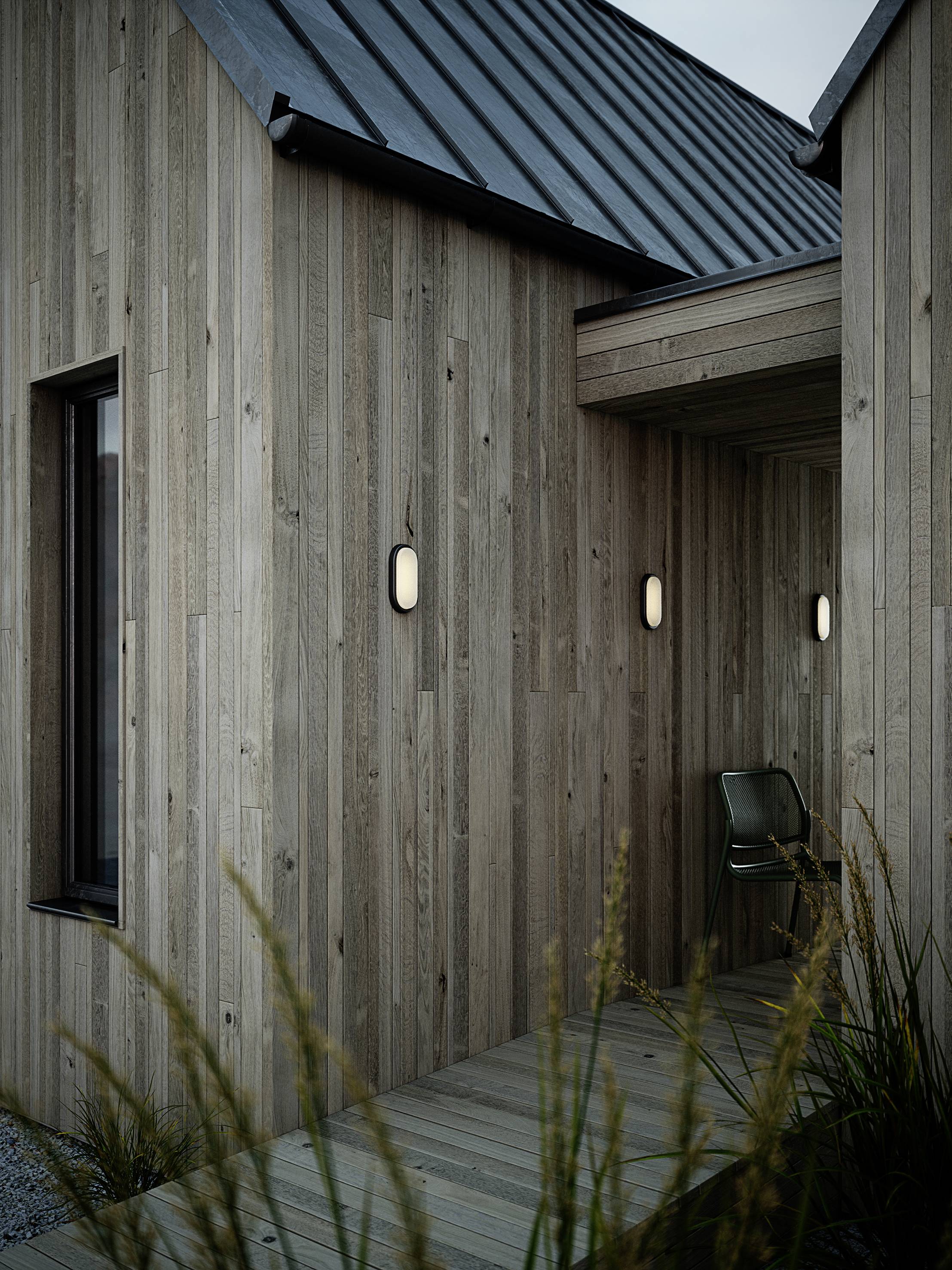 Minimalistisches Holzhaus mit schrägem Metalldach, Außenlampen an den Wänden, ein Stuhl in der Ecke, umgeben von Gräsern.