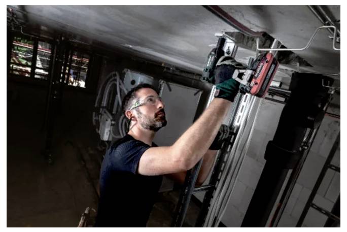 Ein Mann mit Sicherheitsbrille und Handschuhen bedient ein Elektrowerkzeug, während er auf einer Plattform steht und an einer Deckenstruktur in einem industriellen Umfeld arbeitet.