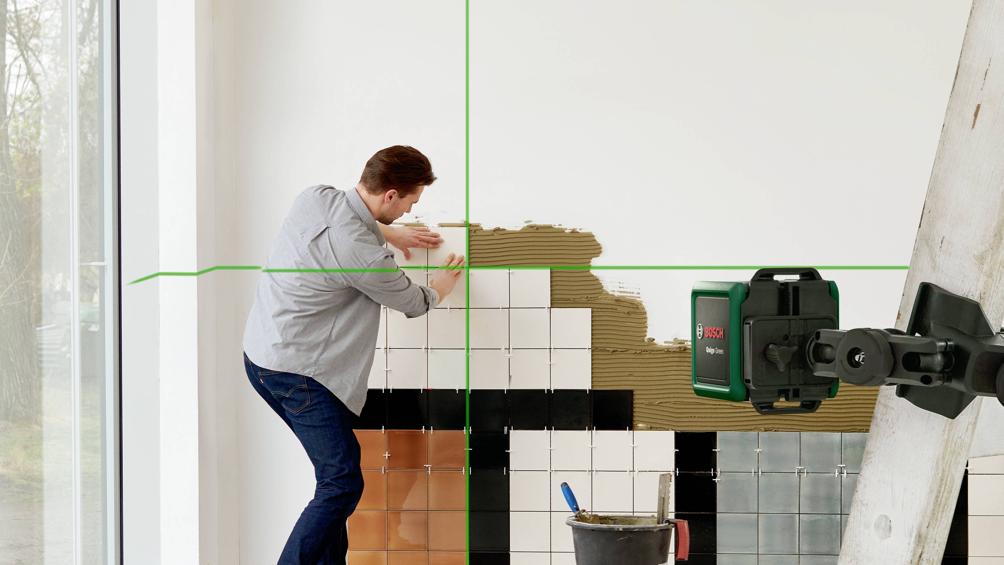 Ein Mann bringt Fliesen an einer Wand an, während ein Laser-Nivelliergerät grüne Linien zur Unterstützung projiziert.