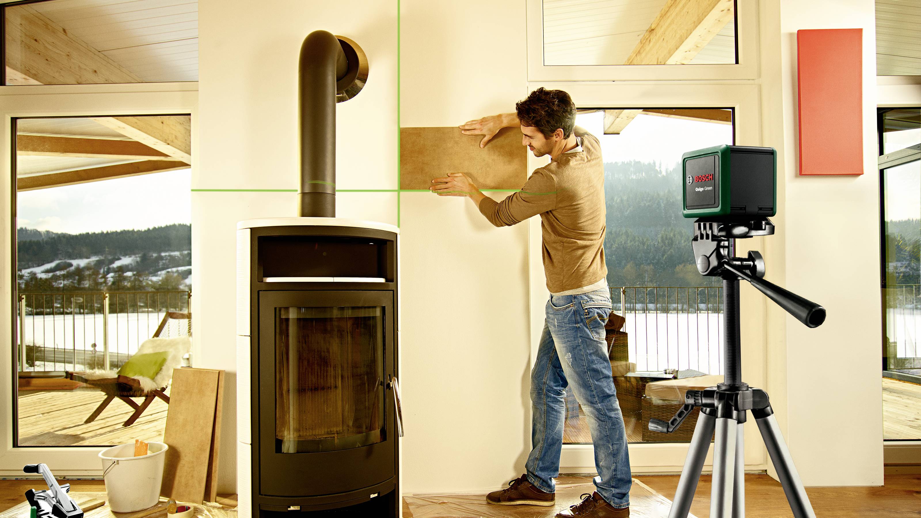 Ein Mann passt ein Holzbrett an eine Wand neben einem Kaminofen an, während ein grüner Laser von einem Gerät auf ein Stativ projiziert wird.