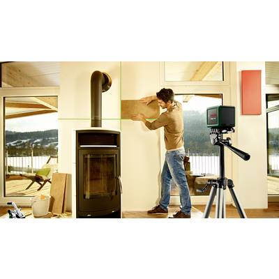 Ein Mann passt ein Holzbrett an eine Wand neben einem Kaminofen an, während ein grüner Laser von einem Gerät auf ein Stativ projiziert wird.