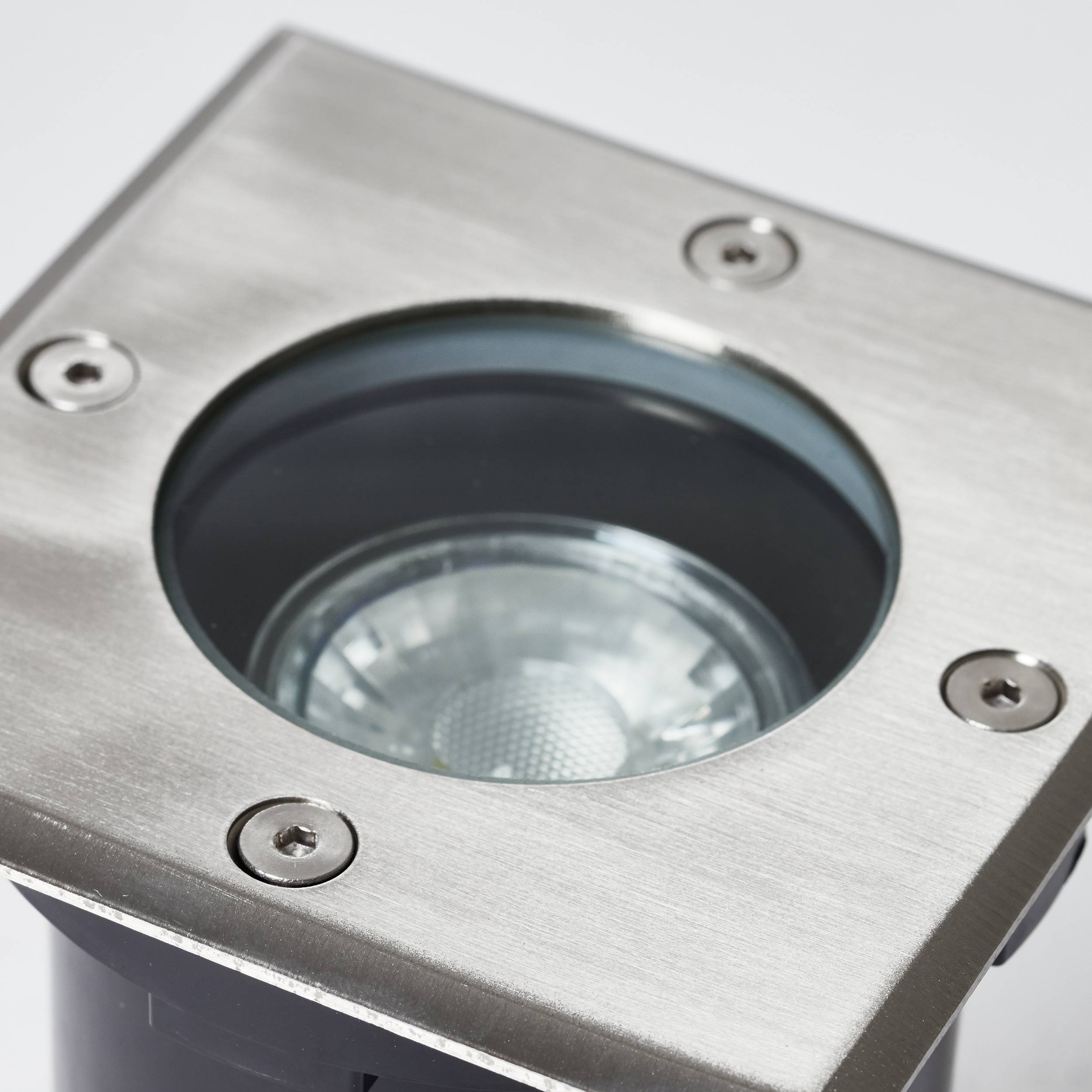 In den Boden eingebauter, quadratischer LED-Strahler aus Edelstahl, umrandet von vier Schrauben. Beleuchtet von oben.