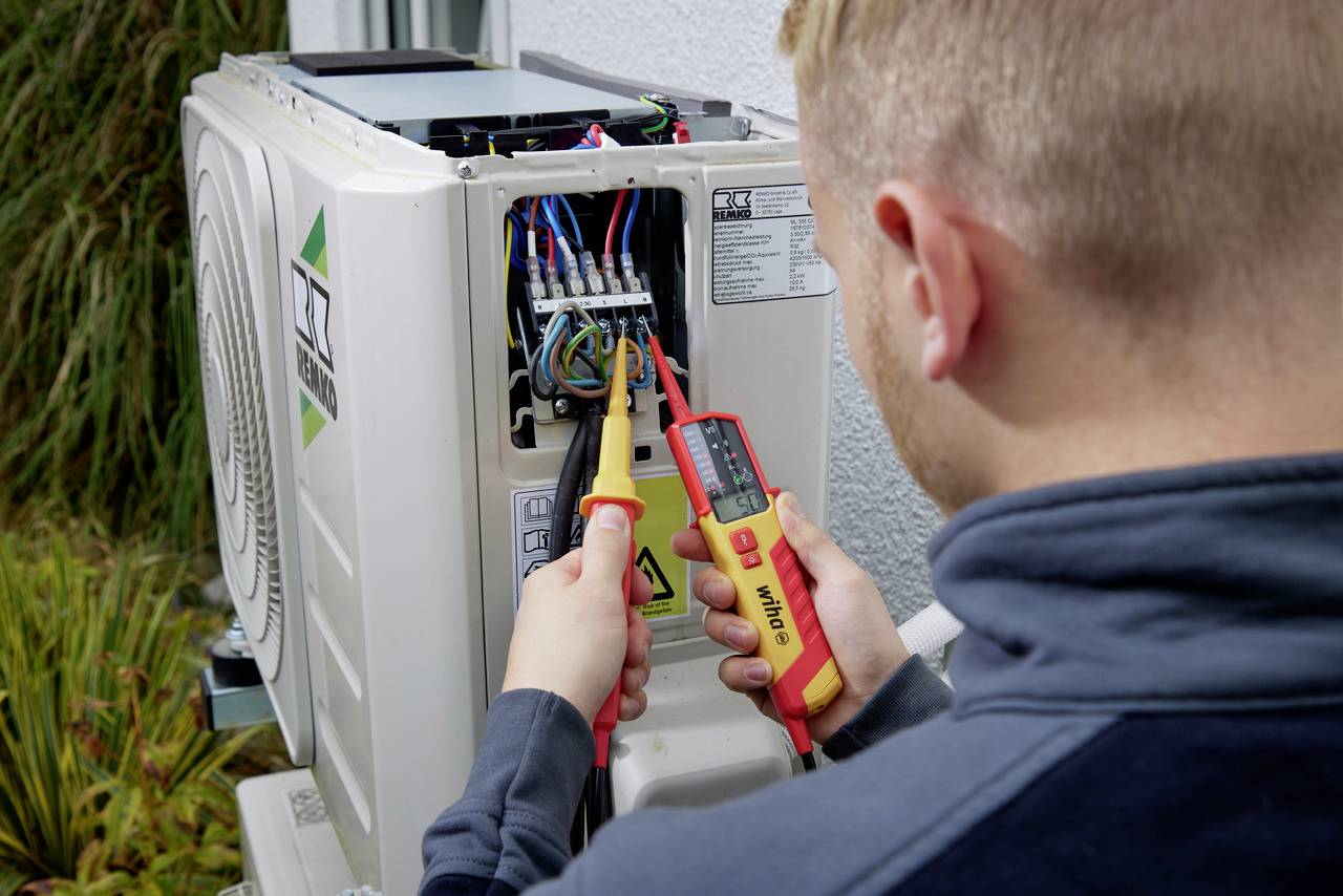 Ein Techniker überprüft mit einem Multimeter die elektrischen Verbindungen in einer Klimaanlage, um die korrekte Spannung sicherzustellen.
