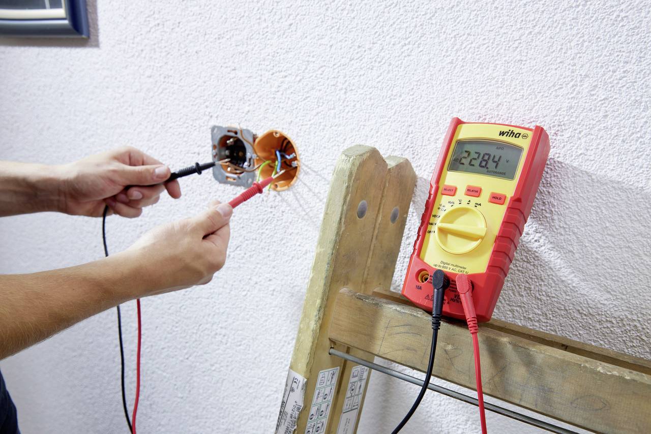 Eine Person misst die Spannung in einer Steckdose mit einem Multimeter, das auf einer Leiter steht und 228,4 V anzeigt.