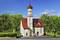 Eine kleine weiße Kirche mit rotem Dach und Turmspitze, umgeben von Bäumen. Menschen stehen vor dem Eingang.