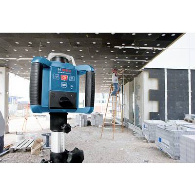 Ein Laser-Nivelliergerät im Vordergrund wird auf einer Baustelle benutzt, während ein Arbeiter im Hintergrund auf einer Leiter arbeitet.