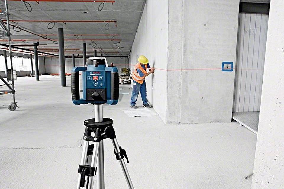 Ein Bauarbeiter misst eine Wand mit einem Laser-Entfernungsmesser in einem unfertigen Raum. Ein Stativ mit Gerät im Vordergrund.