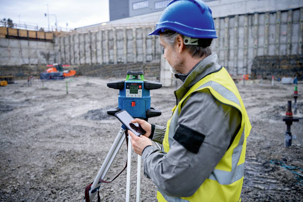 Bauarbeiter in blauer Schutzkleidung bedient ein Vermessungsgerät auf einer Baustelle mit freigelegtem Erdreich im Hintergrund.
