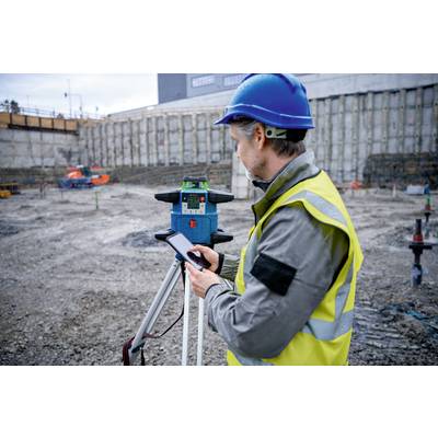 Bauarbeiter in blauer Schutzkleidung bedient ein Vermessungsgerät auf einer Baustelle mit freigelegtem Erdreich im Hintergrund.